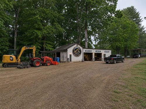 Gravel lot with a yellow excavator, red tractor, small white garage, and black pickup under trees