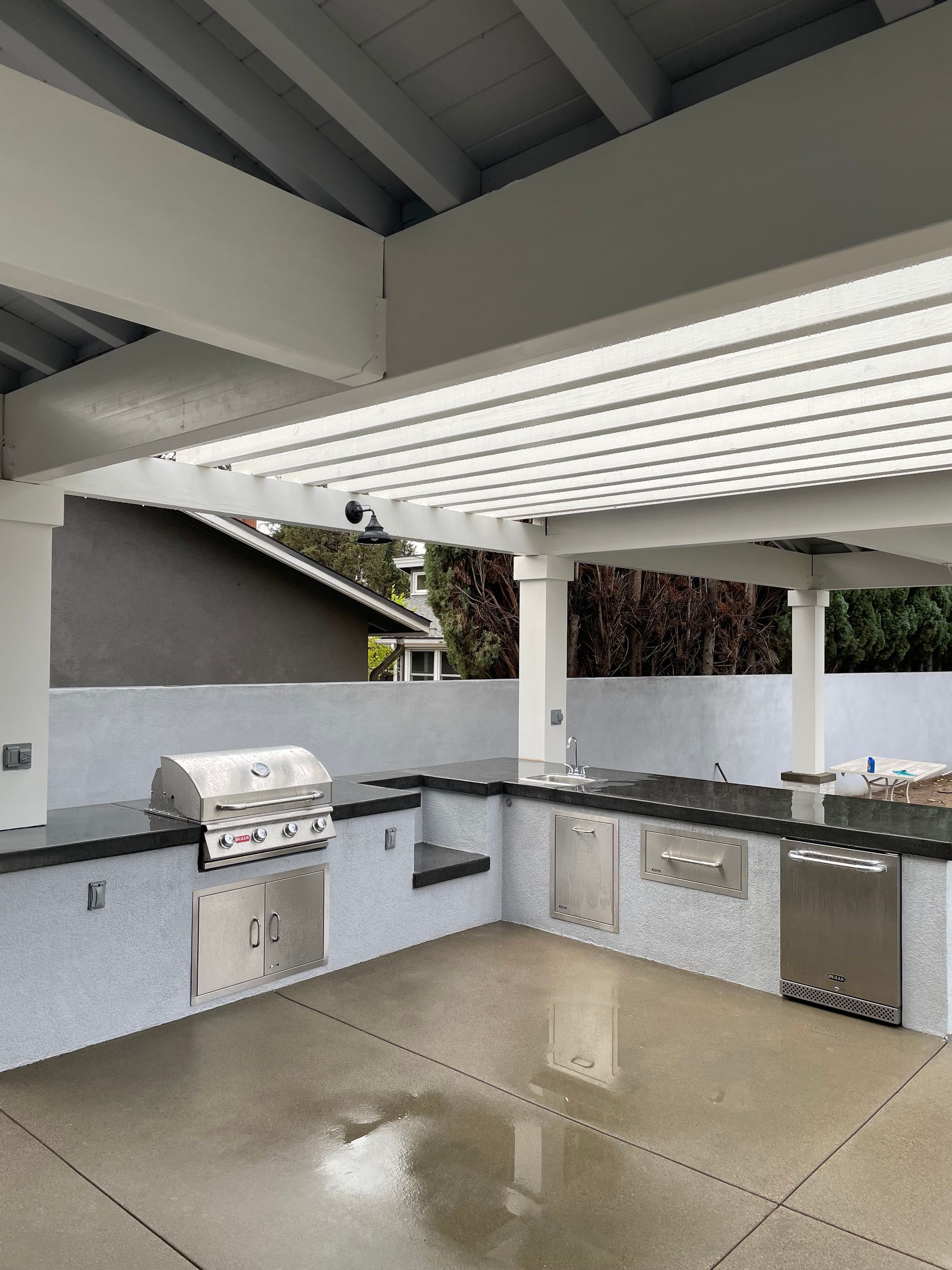 Outdoor kitchen with grill, counters, and cabinetry under a white pergola.