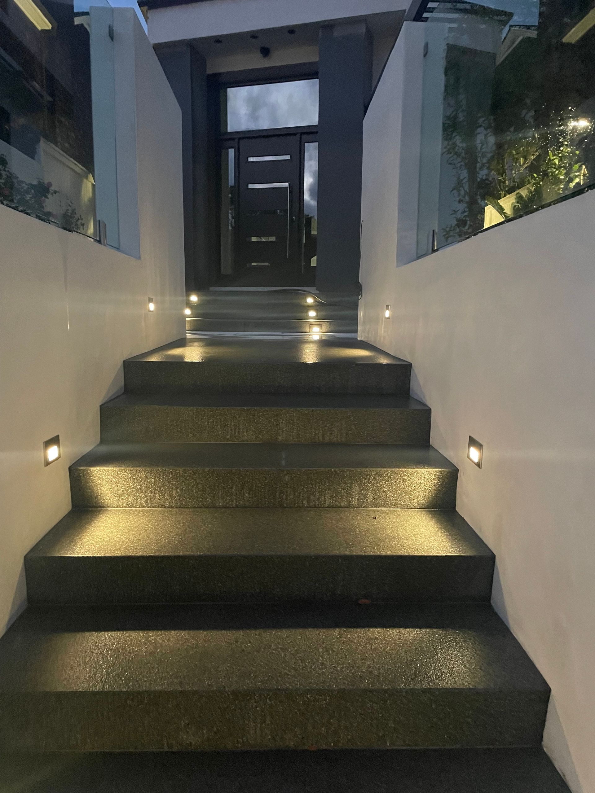 Exterior staircase leading to a door, illuminated by lights on steps and walls; dark steps, white walls.