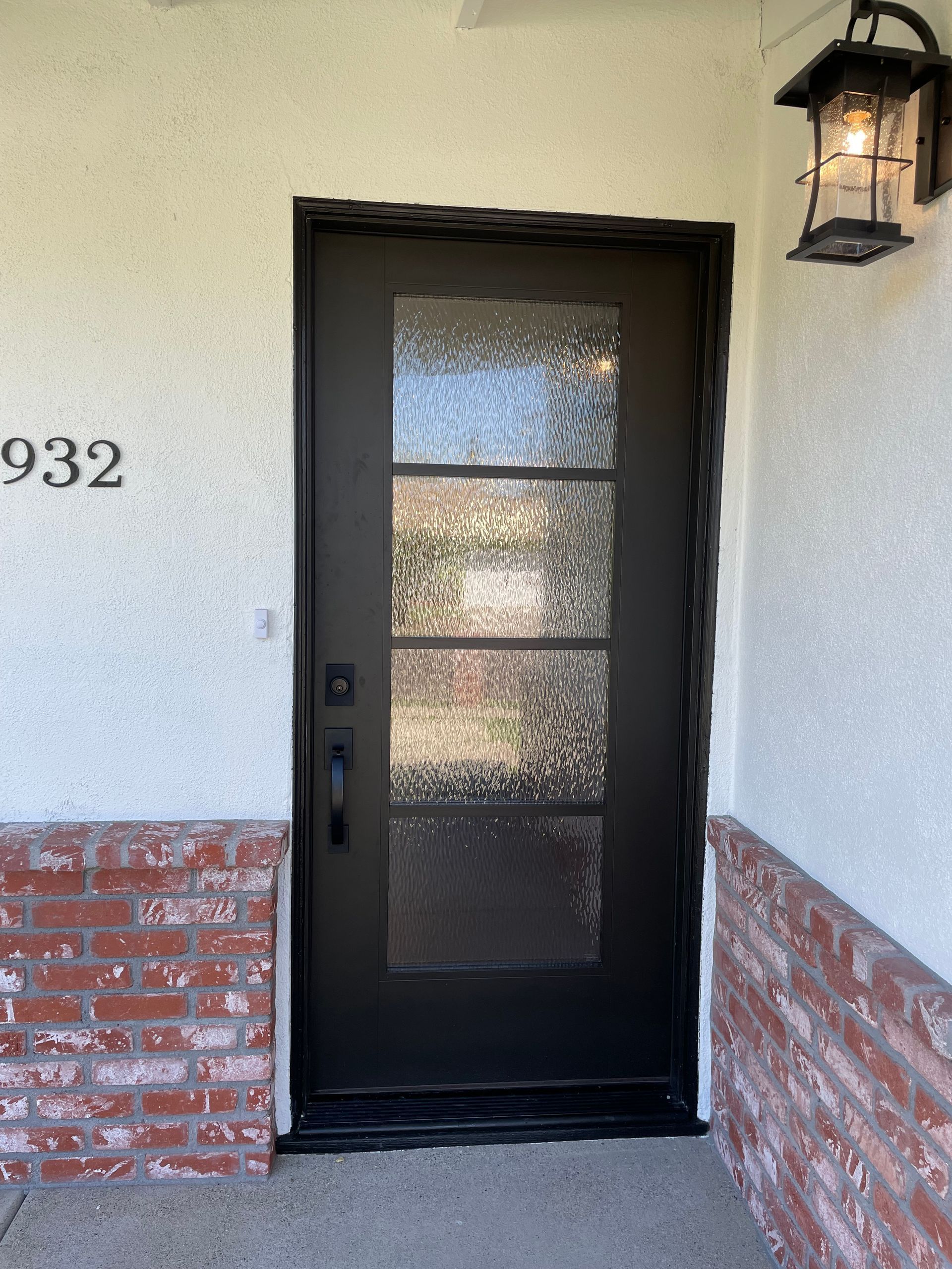 Black front door with three glass panels, set in a white stucco wall with brick base, and a lantern.