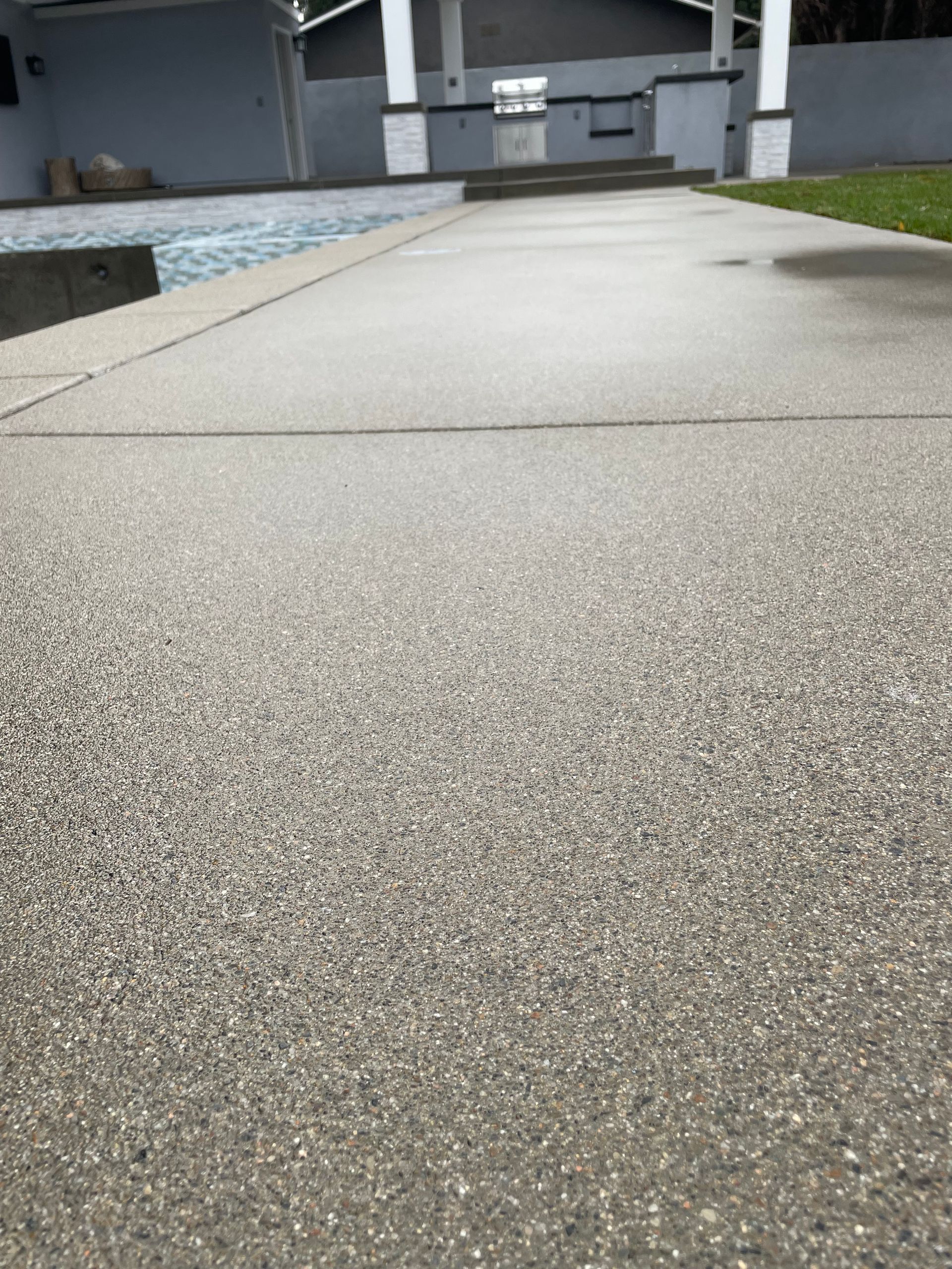 Concrete walkway alongside a swimming pool, leading towards a covered structure; overcast conditions.