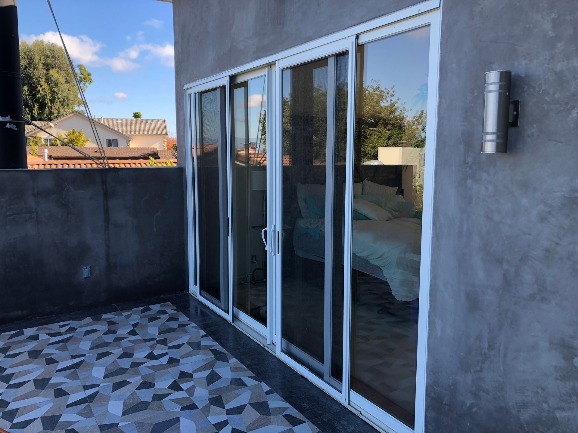 Sliding glass doors on a patio with patterned tile flooring, a gray stucco wall, and a mounted light fixture.