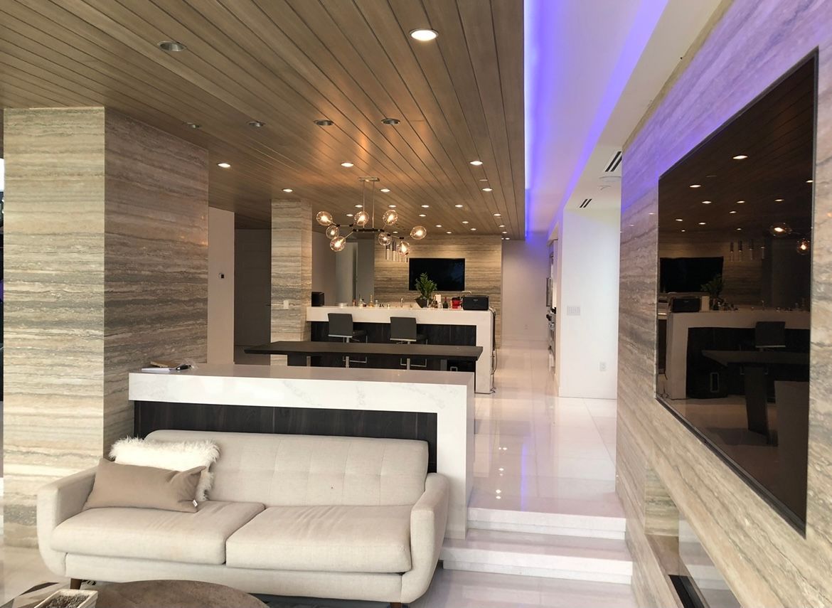 Modern interior with neutral tones: living room, kitchen, and dining area. Beige sofa, marble walls, wooden ceiling, and ambient lighting.