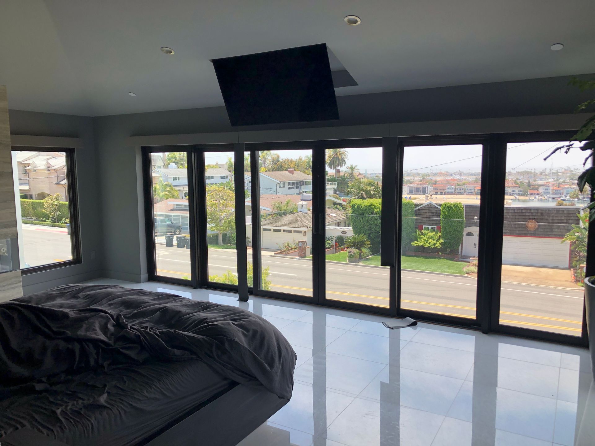 Bedroom with bed, large windows overlooking a neighborhood, and a TV mounted on the ceiling.
