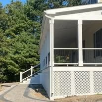 A white porch with lattice skirting and a metal railing sits beside a paved walkway, framed by trees on a sunny day.