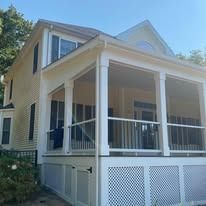 A two-story, yellow-sided house featuring a spacious, white-framed screened porch with lattice skirting.
