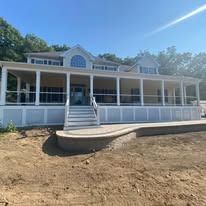 A large, beige house with a wrap-around porch and white railings stands above a stone patio and unfinished dirt yard.