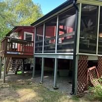 A tan screened-in porch on stilts sits next to a smaller, elevated red deck with stairs in a wooded backyard.