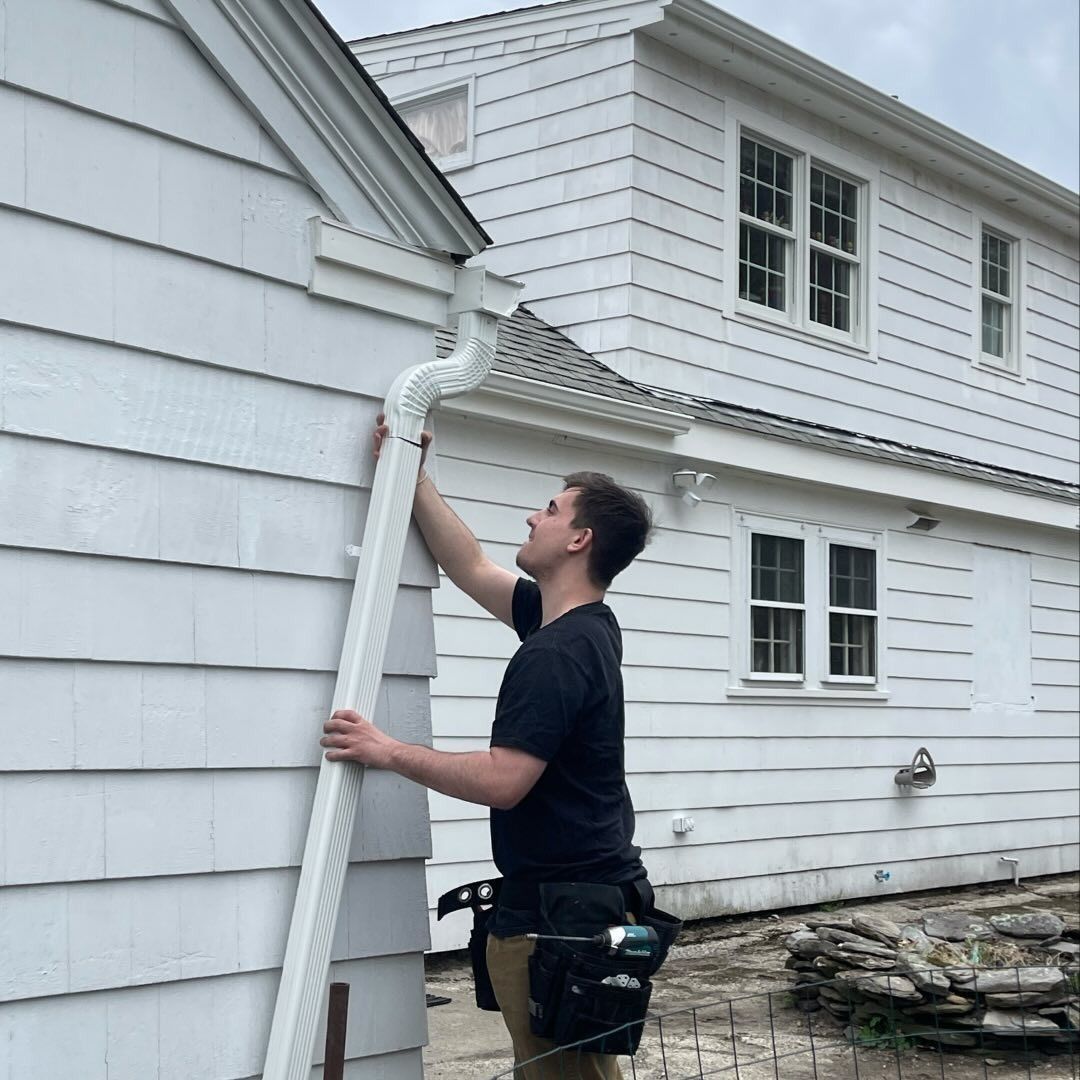 A person wearing a tool belt installs a white downspout on the side of a white shingled house.