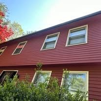 The side of a two-story house with red horizontal siding and several white-framed windows above a green hedge.