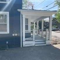 A blue house with a white front porch featuring stairs and railings, seen from the side on a paved street.