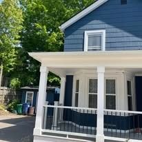 A blue two-story house with a white wrap-around porch, columns, a railing, and a bay window, next to a small shed.