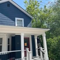 A blue two-story house with a white front porch and red door. A person on a ladder is working near the roofline.