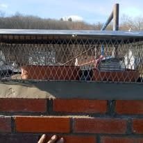 A close-up of a brick chimney top, featuring a metal mesh screen cover and a chimney cap against a clear blue sky.