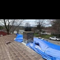 A brick chimney stands on a brown shingled roof, which is partially covered by a large blue tarp.