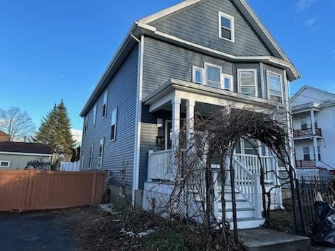 A blue, two-story house with white trim, a front porch, and a bare arbor in the yard under a clear blue sky.
