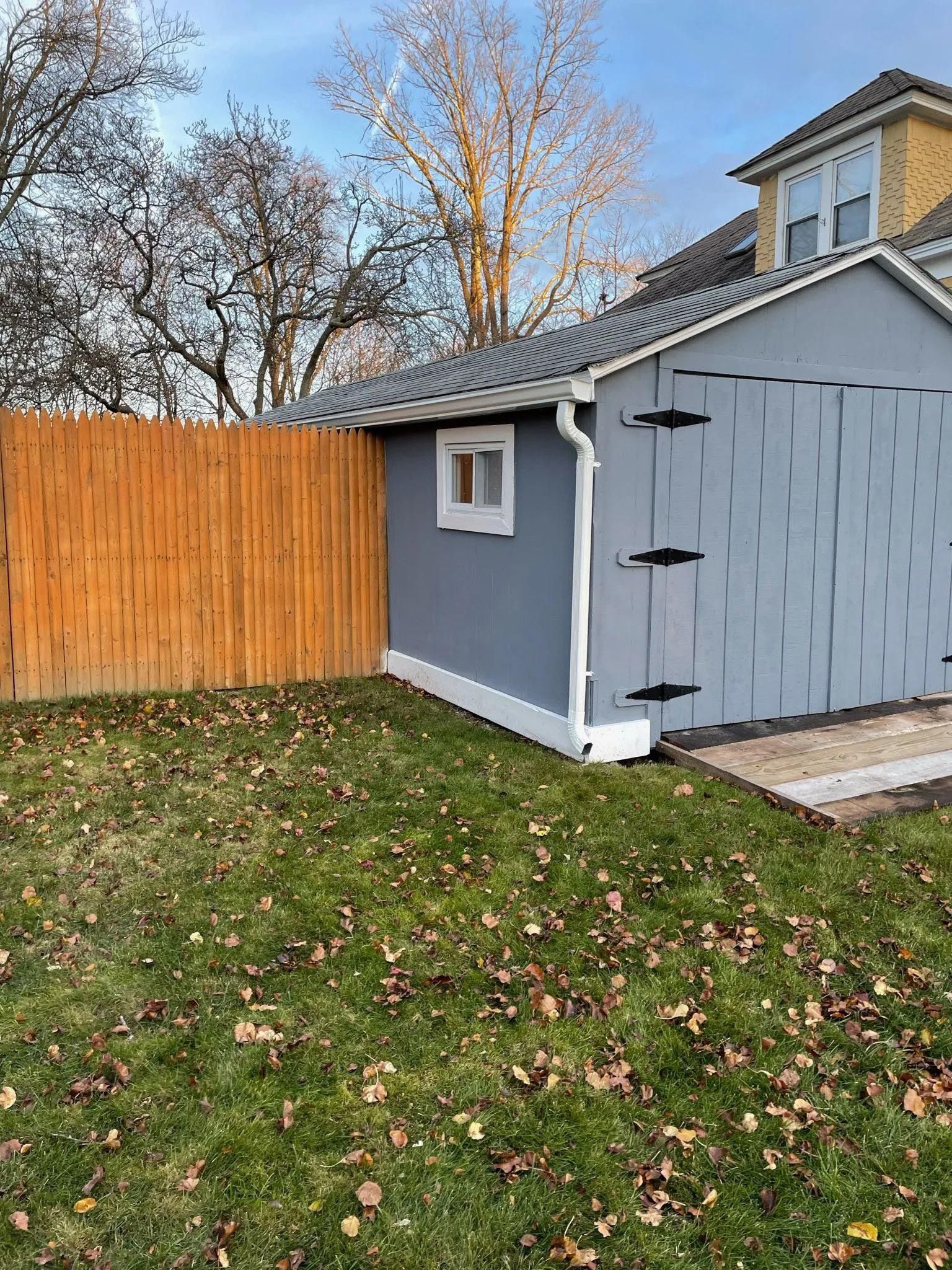 A grey shed with a white trim and gutter stands in a grassy yard next to a tall, orange wooden fence.
