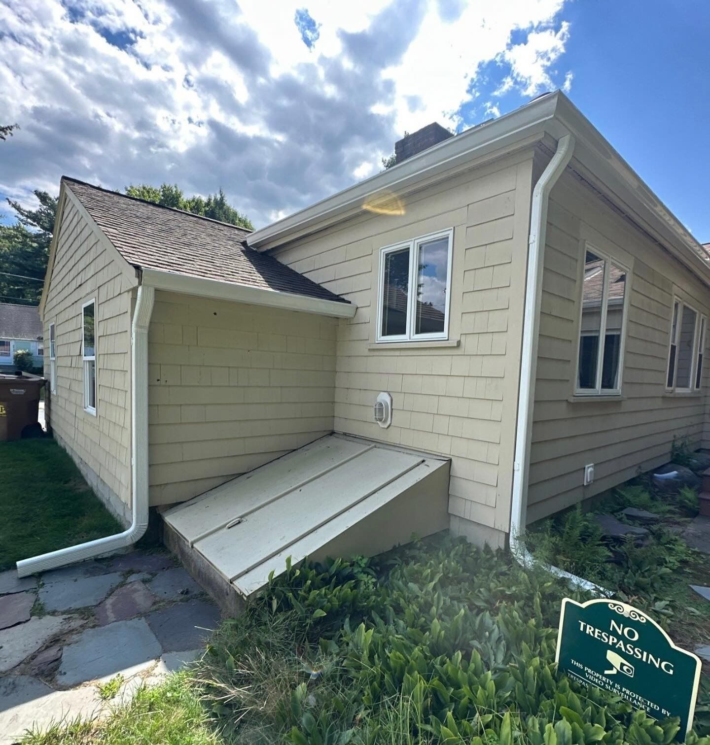 Side view of a beige house with an exterior basement bulkhead door, white trim, and a small no-trespassing sign.