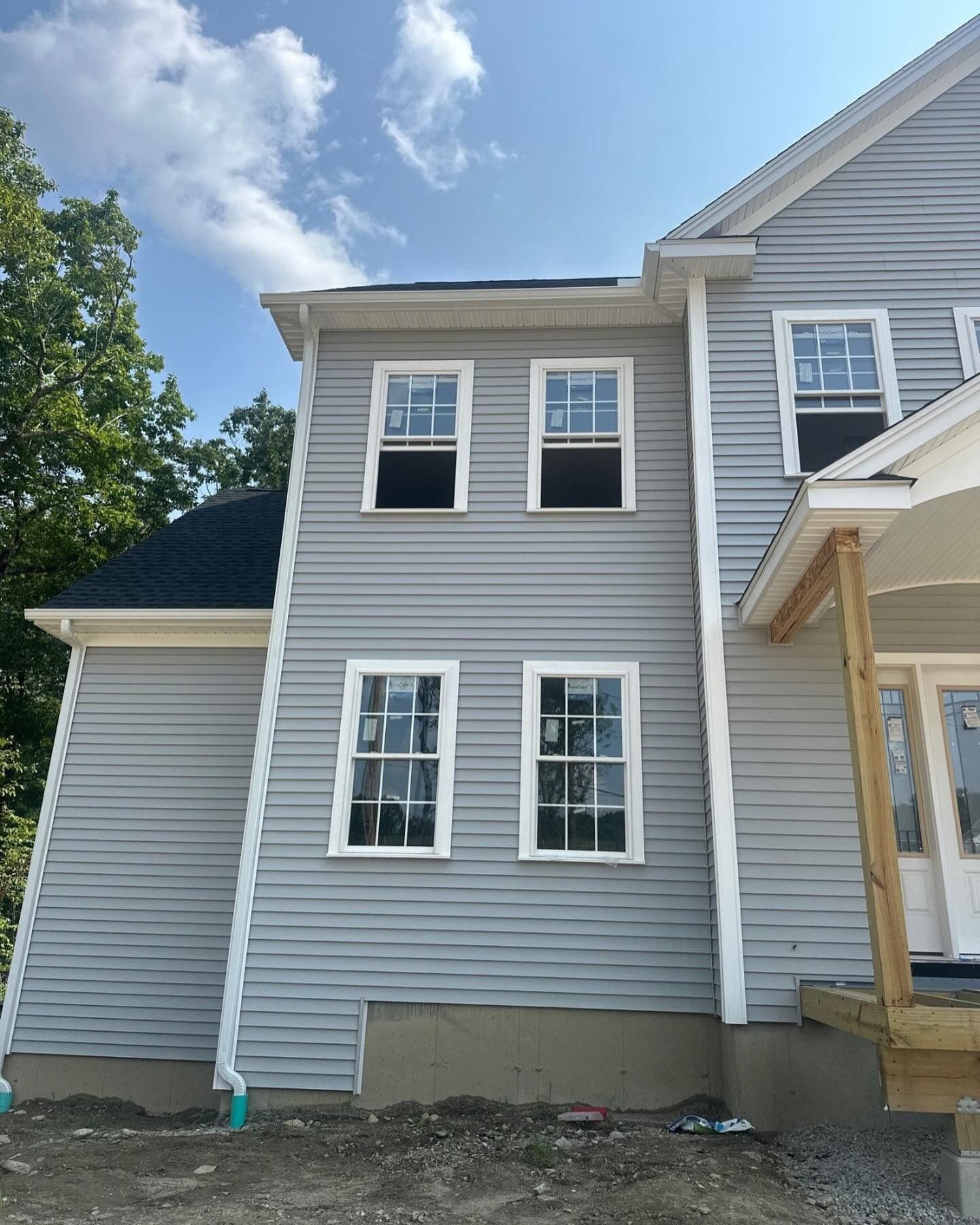 A gray, two-story house under construction with white window frames and a partial front porch against a blue sky.