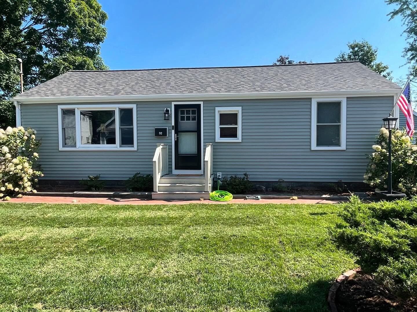 A light blue ranch-style house with gray roof shingles, a front door with a storm door, and green lawn on a sunny day.