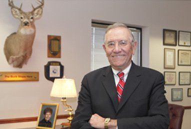 Man in suit, arms crossed, smiles in office with deer head mount and diplomas.
