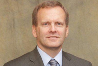 Man in a suit smiles, against a neutral background.