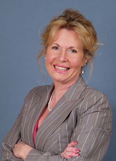 Woman in gray pinstripe suit, arms crossed, smiling, pearl necklace.