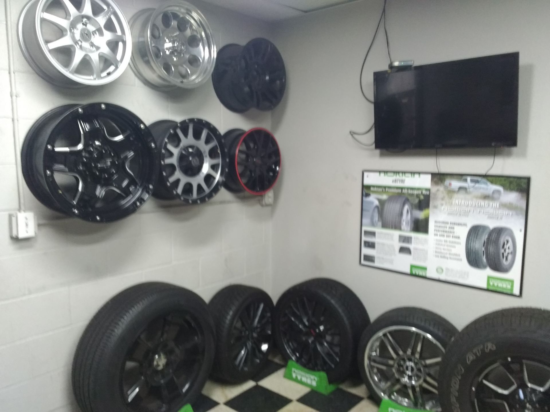 Wheels and tires displayed on a wall and floor in an auto shop. Various styles and colors. A TV and advertising sign are also visible.