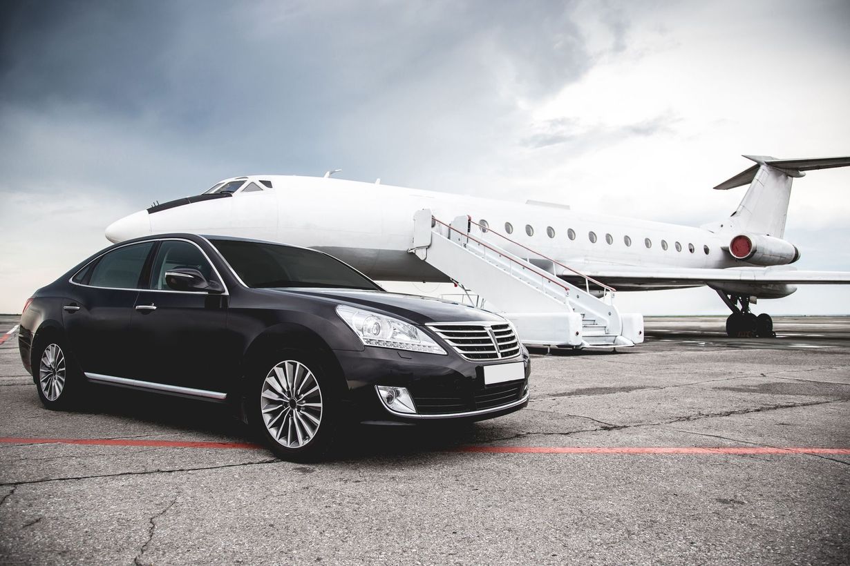 A black sedan parked on a runway in front of a white private jet with a cloudy sky.