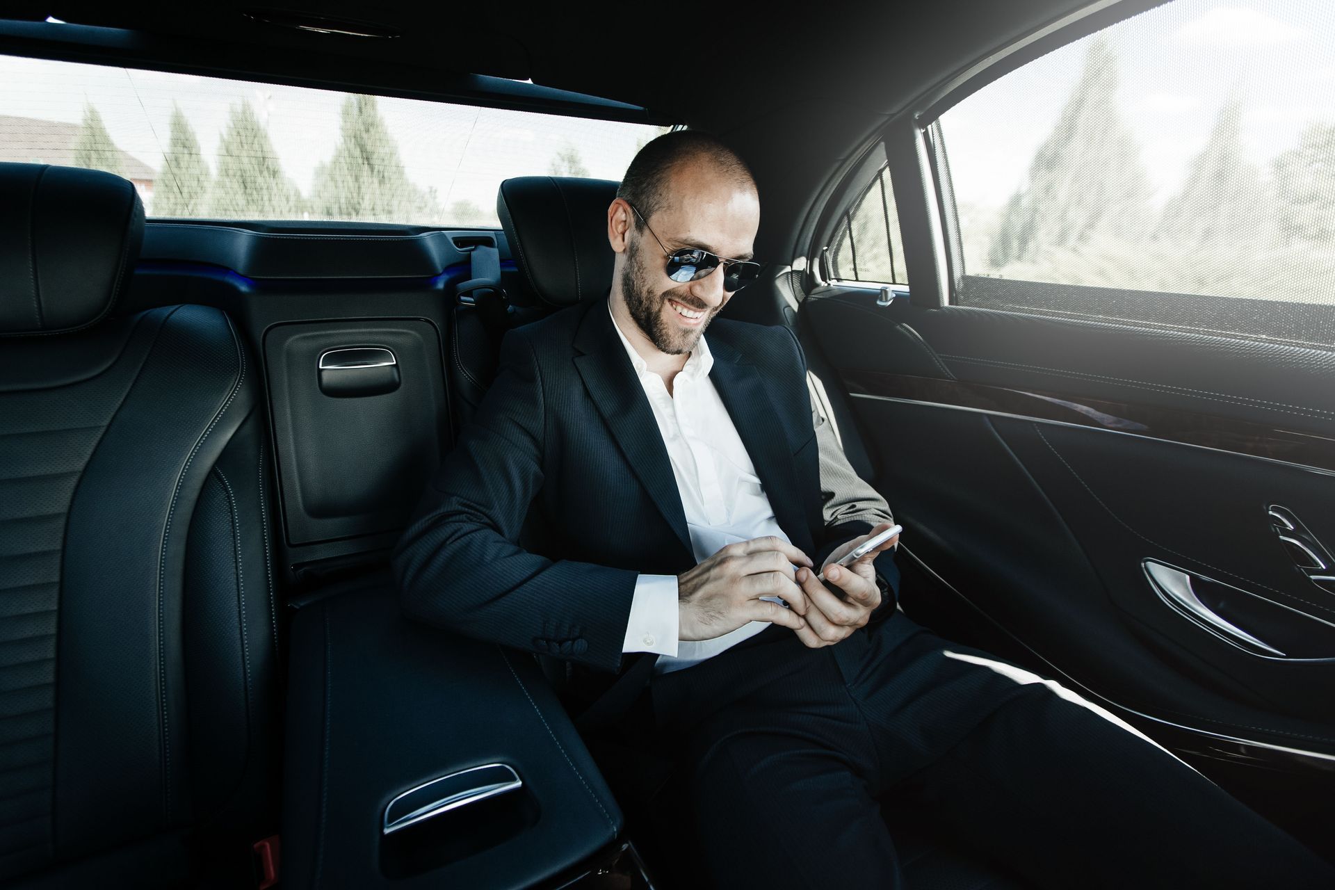 Man in suit smiling while looking at phone in the backseat of a luxury car.