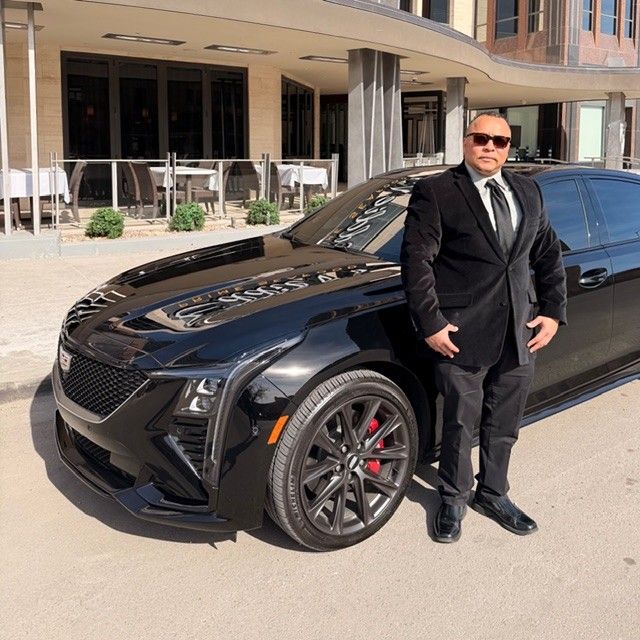 Man in a black suit standing next to a black Cadillac, in front of a building with outdoor seating.