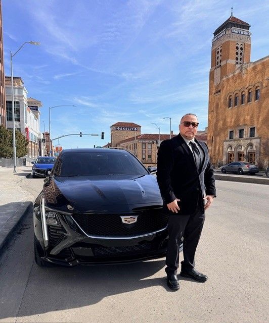Man in suit standing next to a black Cadillac on a city street with a church in the background.
