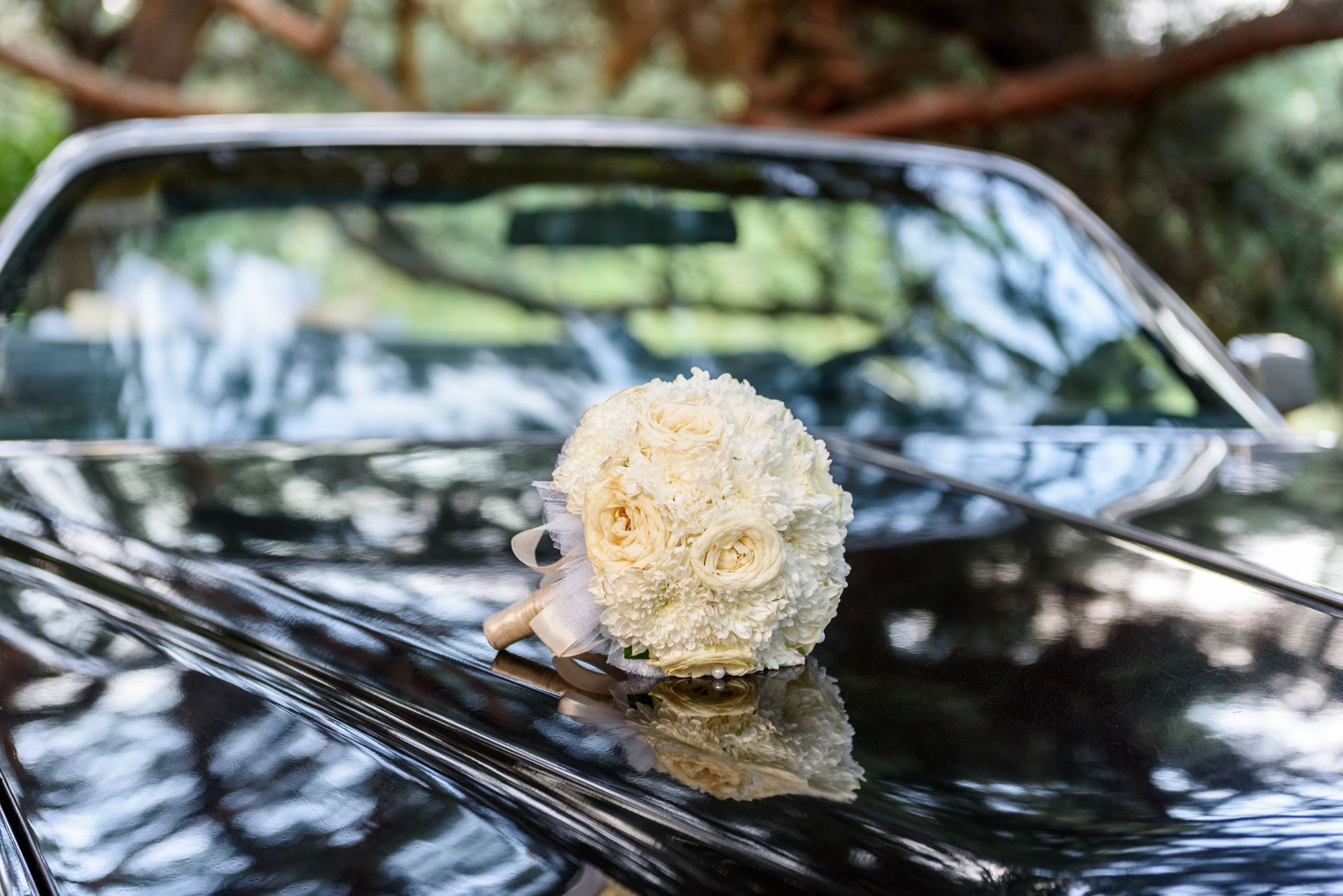Bouquet of white flowers on the hood of a shiny black car, with greenery in the background.