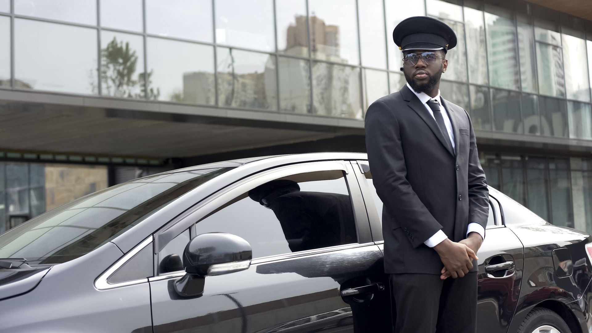 A person in a black chauffeur uniform stands beside a black car in front of a modern building.