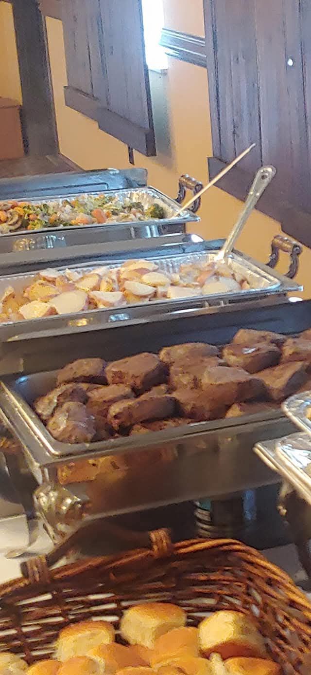 Buffet with food in serving trays: vegetables, potatoes, and meat. A basket of rolls is in the foreground.