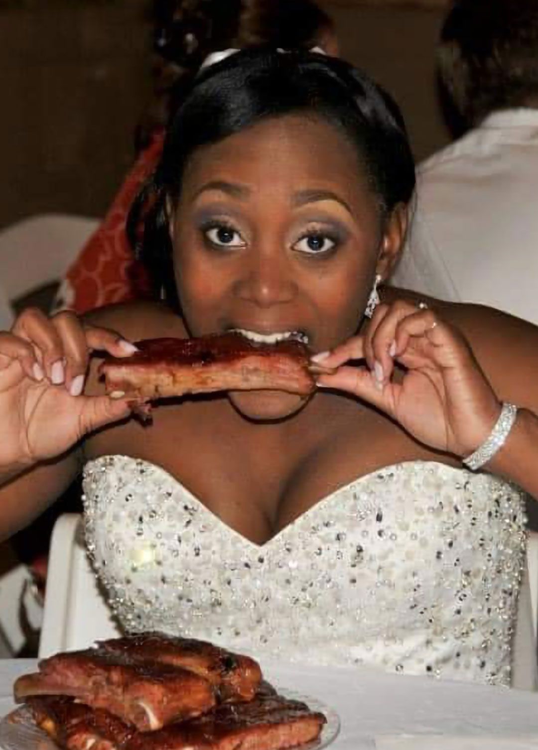Bride in a white gown eating ribs at a table; a plate of ribs in front of her.
