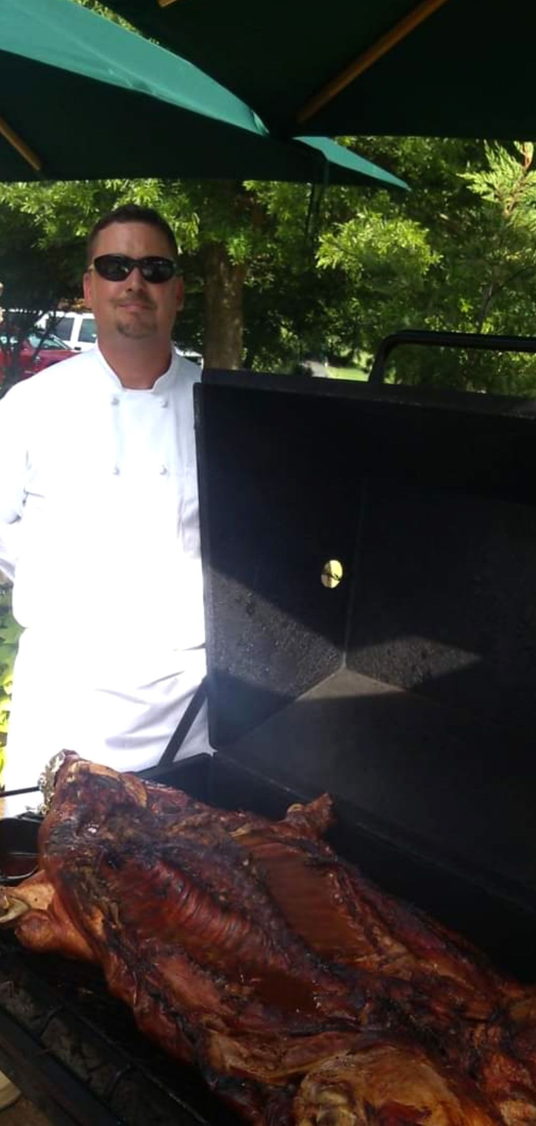 Chef wearing sunglasses stands near a smoker with a roasted pig; outdoor setting.