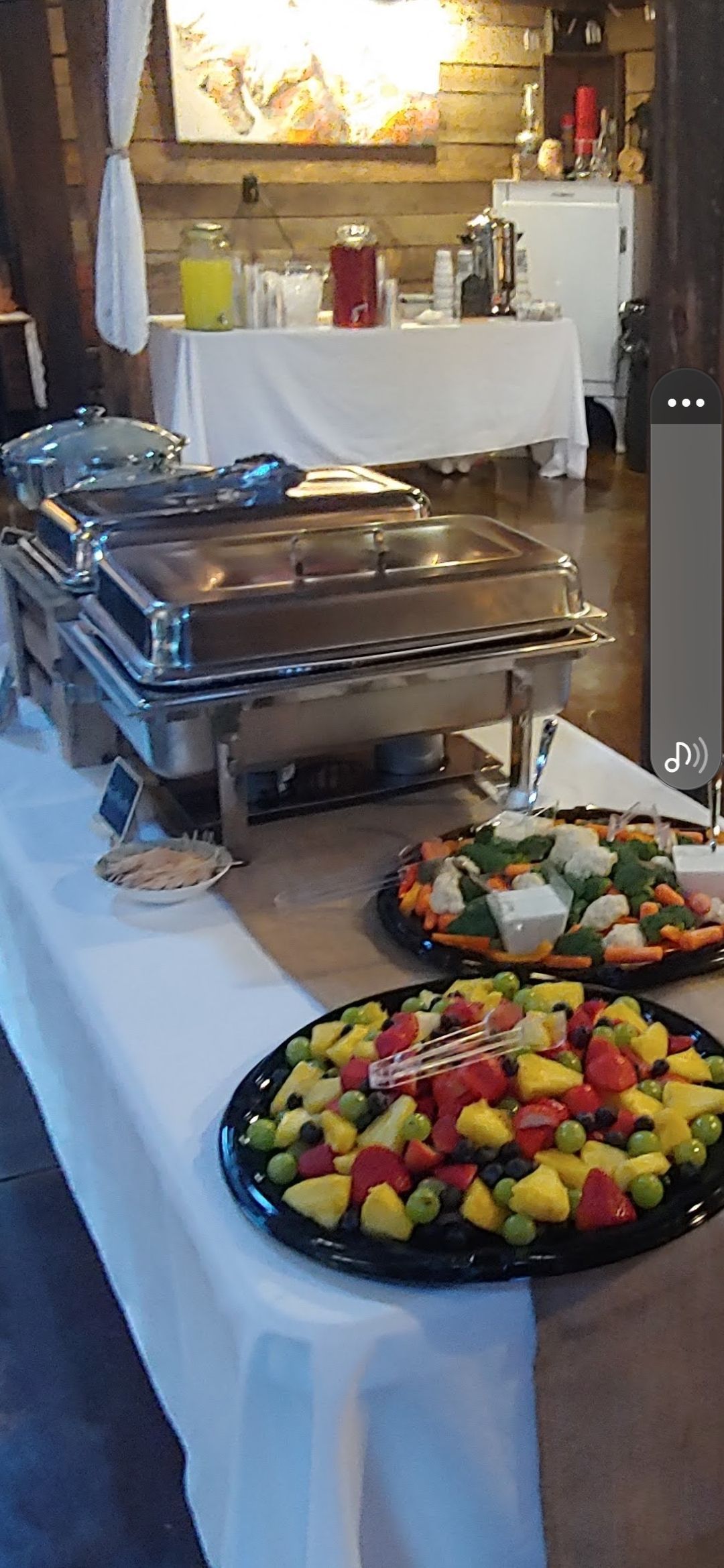 Buffet table with chafing dishes, fruit platters, and beverage station in a rustic setting.