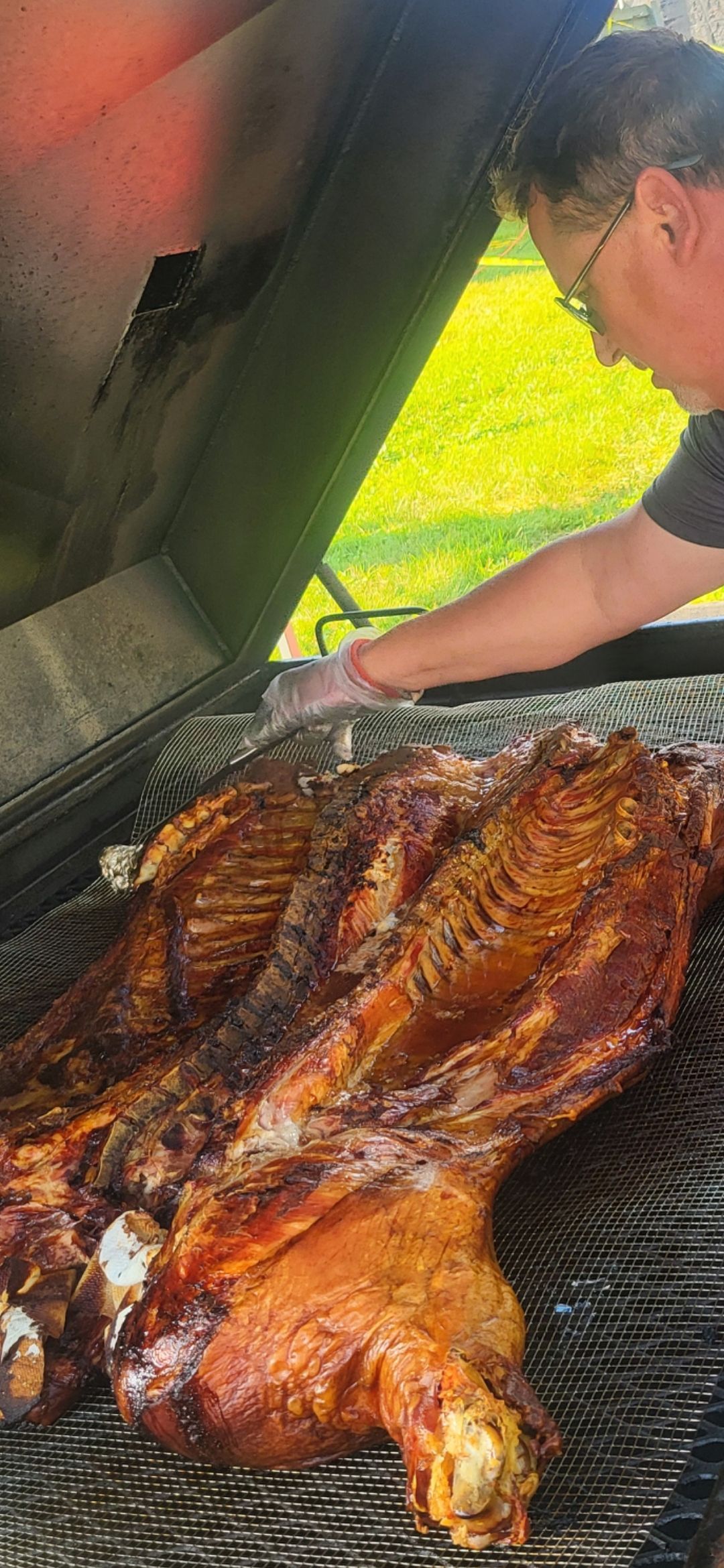 A whole roasted animal on a grill. A person in glasses, wearing gloves, tends to the meat.