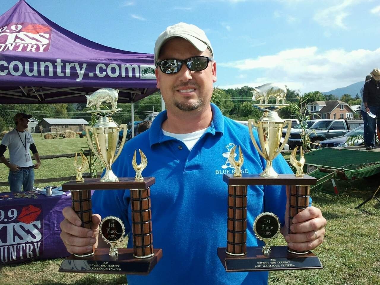 Man in blue shirt, holding two trophies, in front of a tent and a sunny outdoor scene.