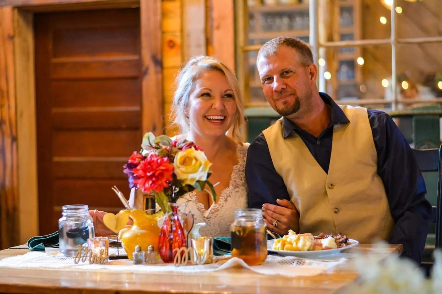 Bride and groom at wedding reception, smiling, seated at a table with flowers and food. Rustic setting.