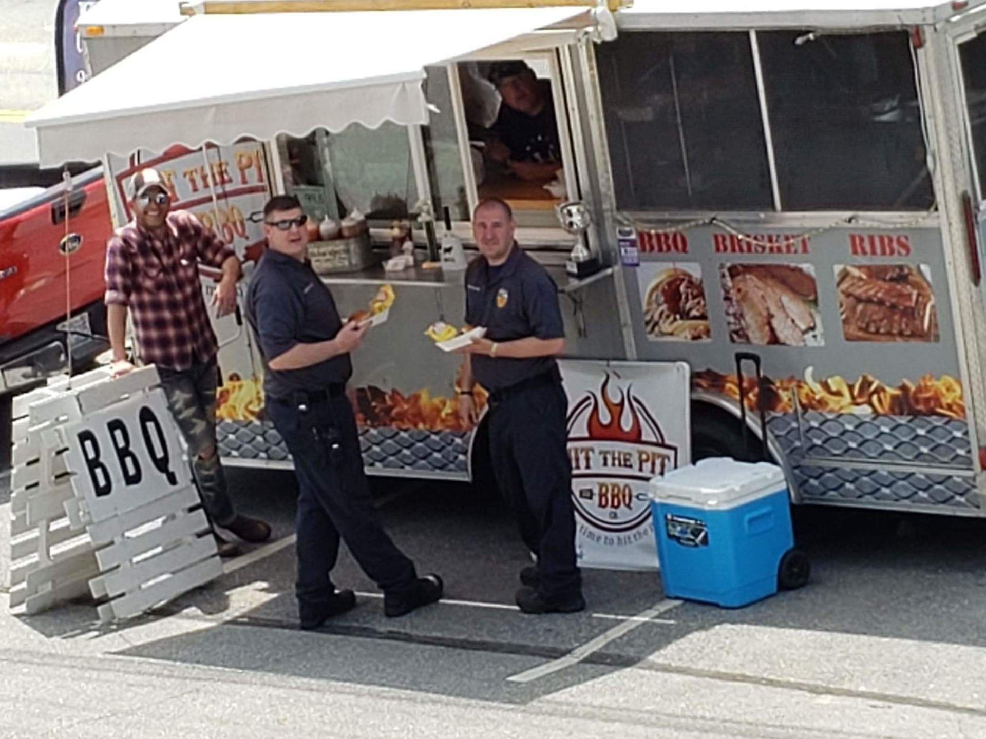 Two uniformed men and a man in a plaid shirt pose near a BBQ food truck, holding food.