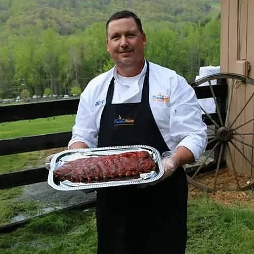 Chef holding a tray of ribs outdoors near a fence and a wooden wagon wheel.