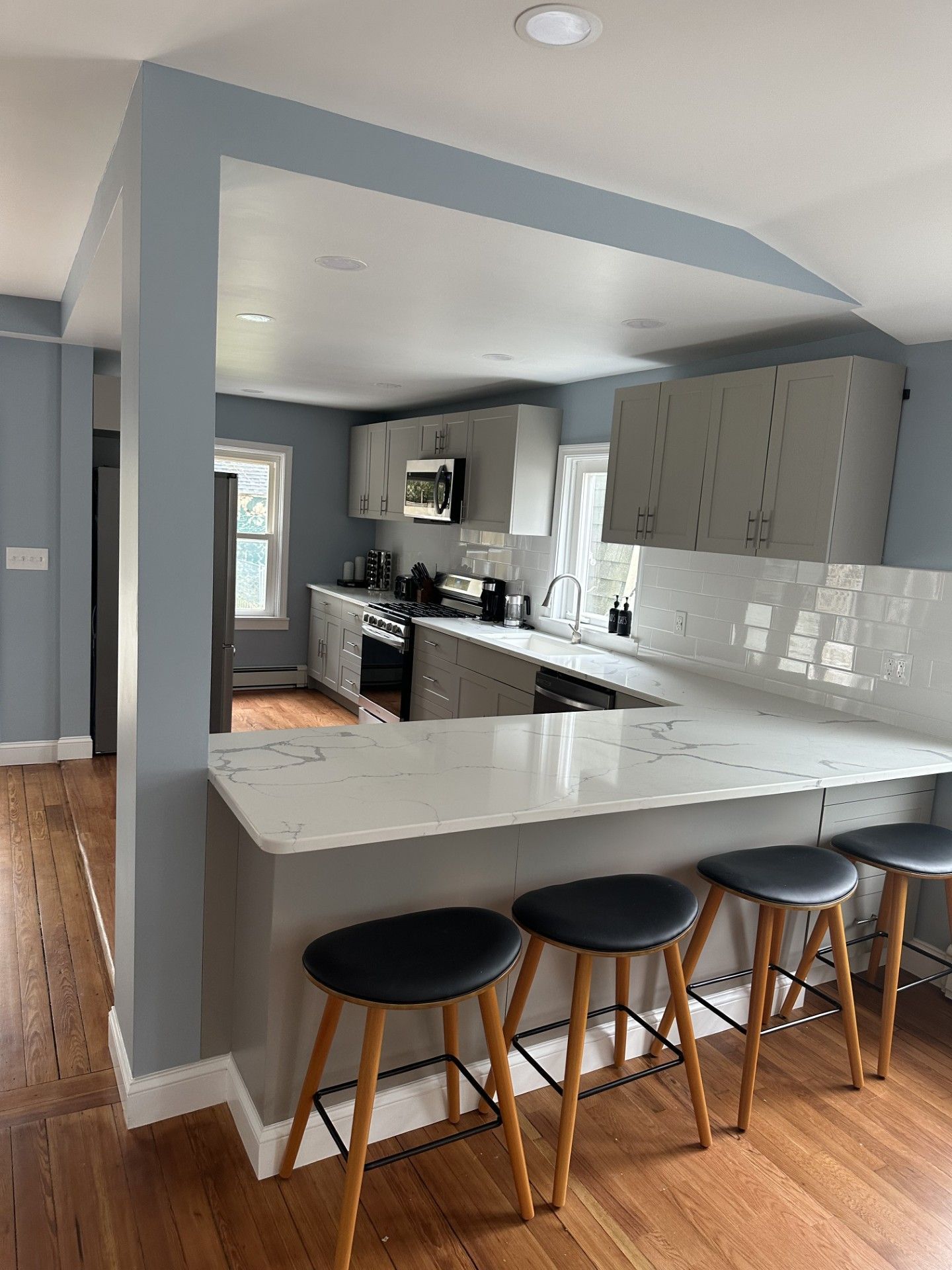 Kitchen with a breakfast bar and gray cabinets, with wood floors. Three bar stools sit at the counter.