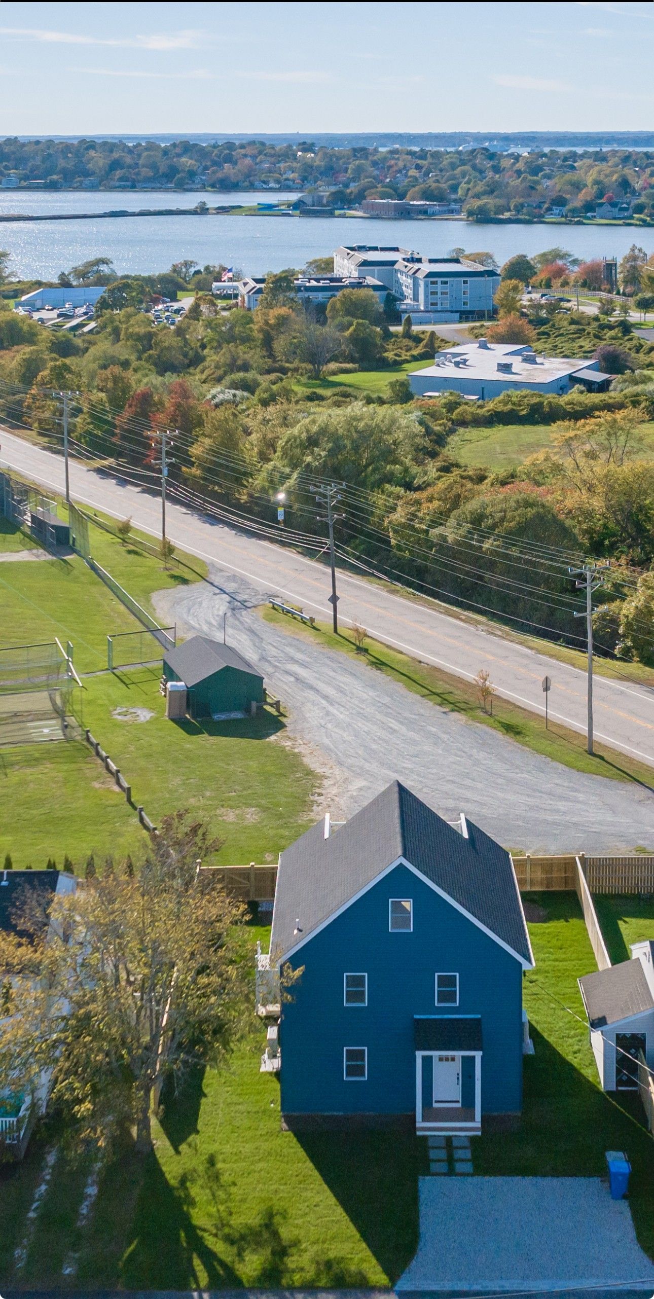 Aerial view of a blue house with a dark roof near a road, overlooking a body of water and buildings.