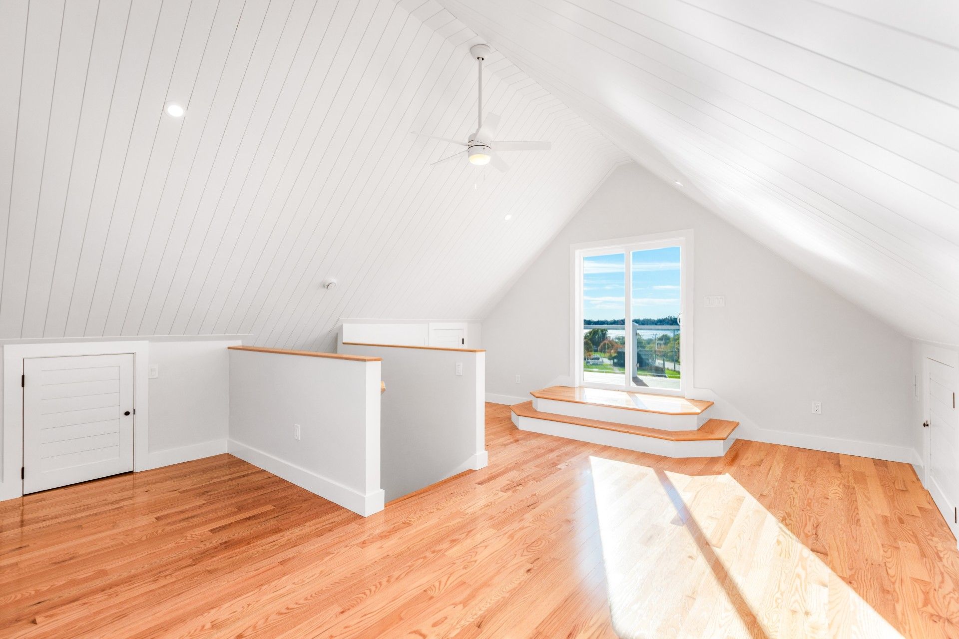 Bright, white attic room with wooden floors, sloped ceiling, and large window overlooking a body of water.
