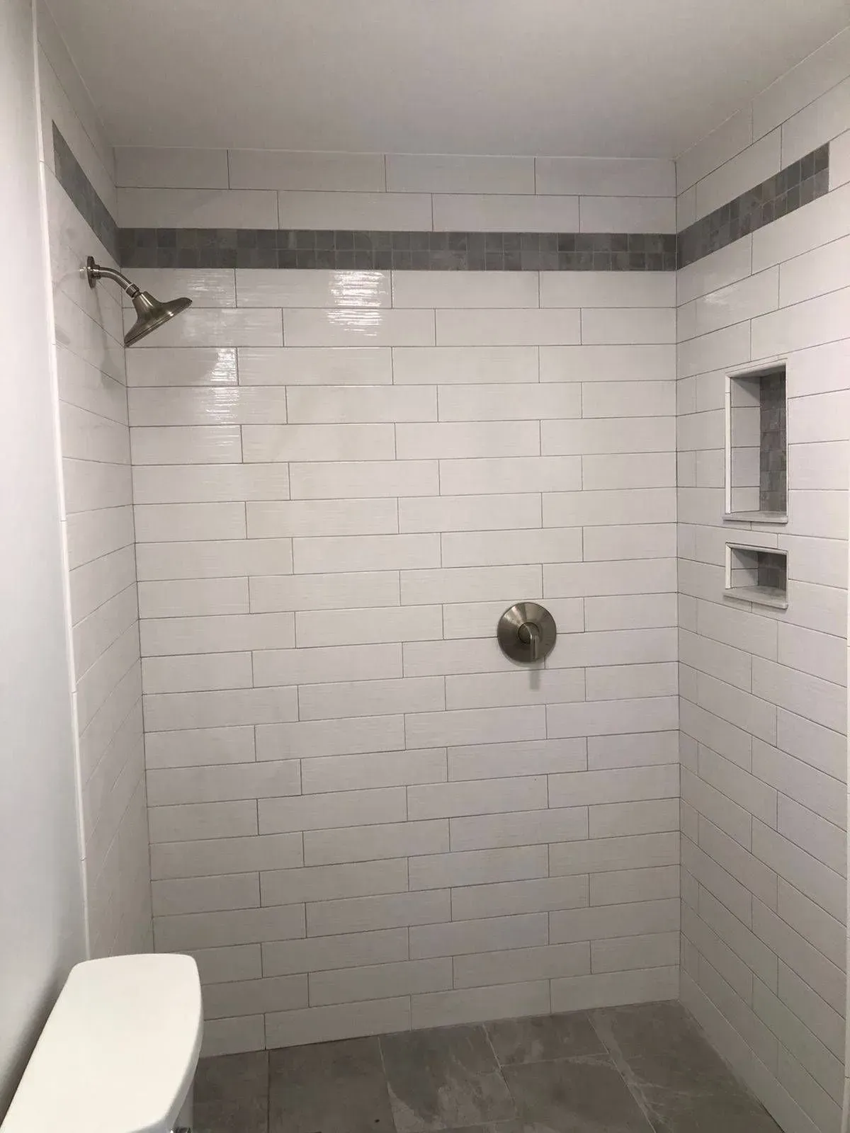 Tiled shower stall with white rectangular tiles, gray accent stripe, built-in shelf, and rain shower head.