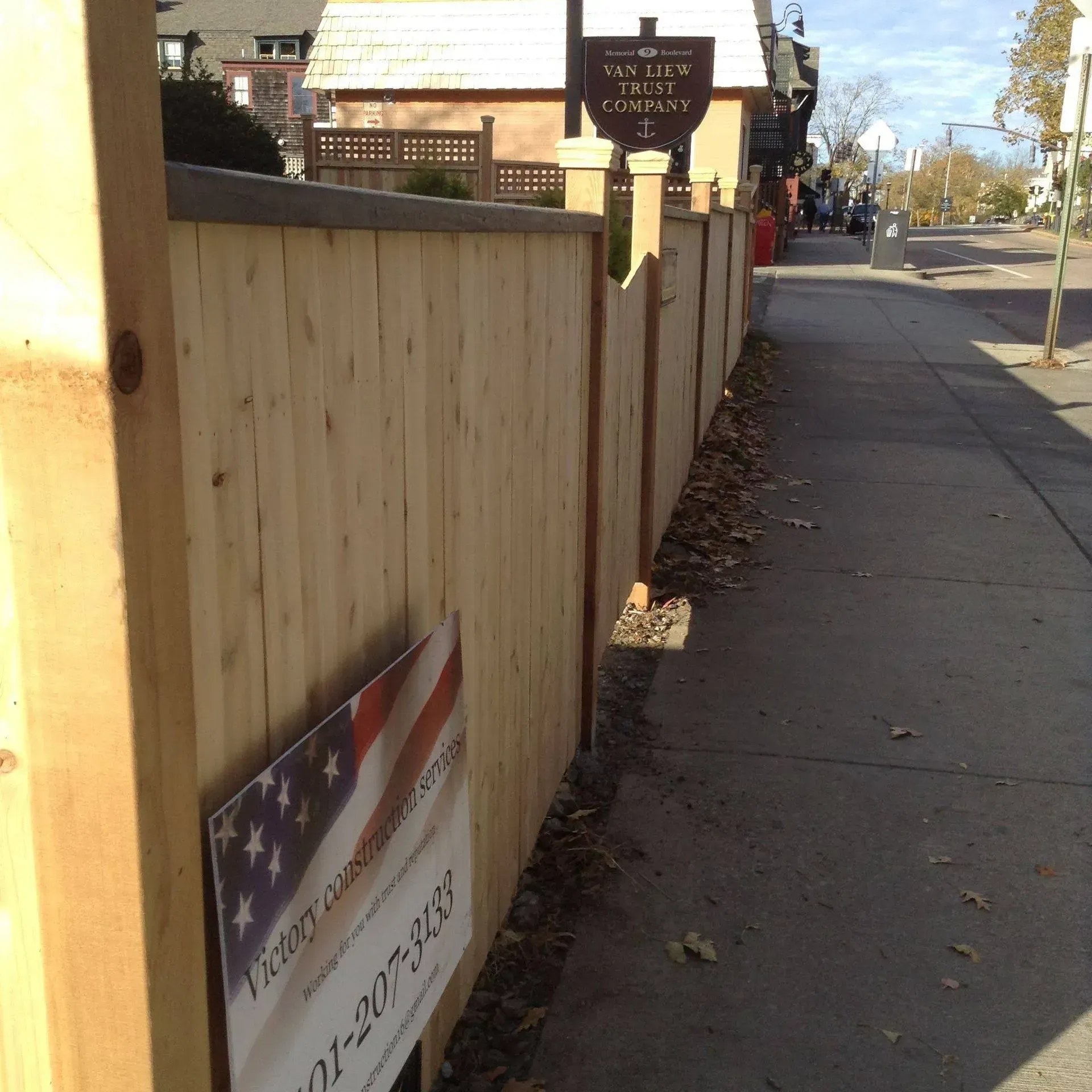 Wooden fence bordering a sidewalk, with a sign for a business and a street in the background.