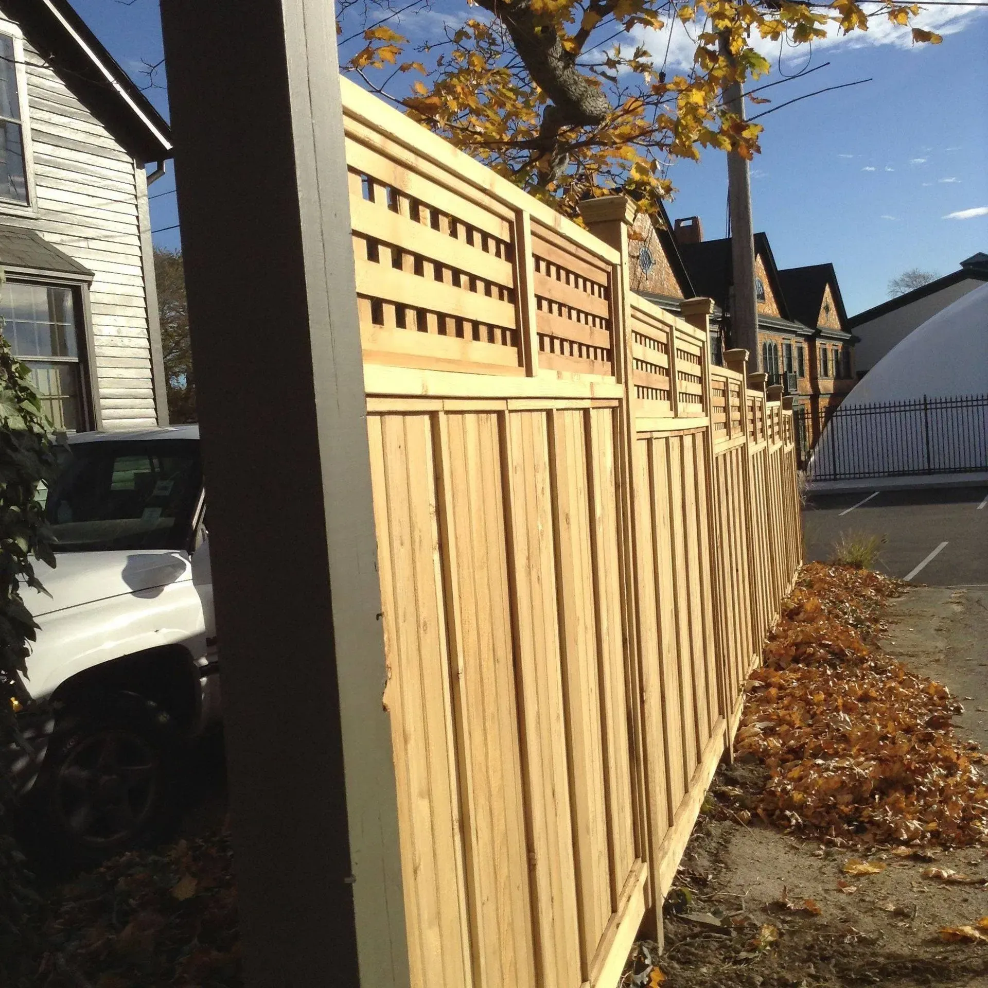Wooden fence with lattice top running along a street, fallen leaves, blue sky.