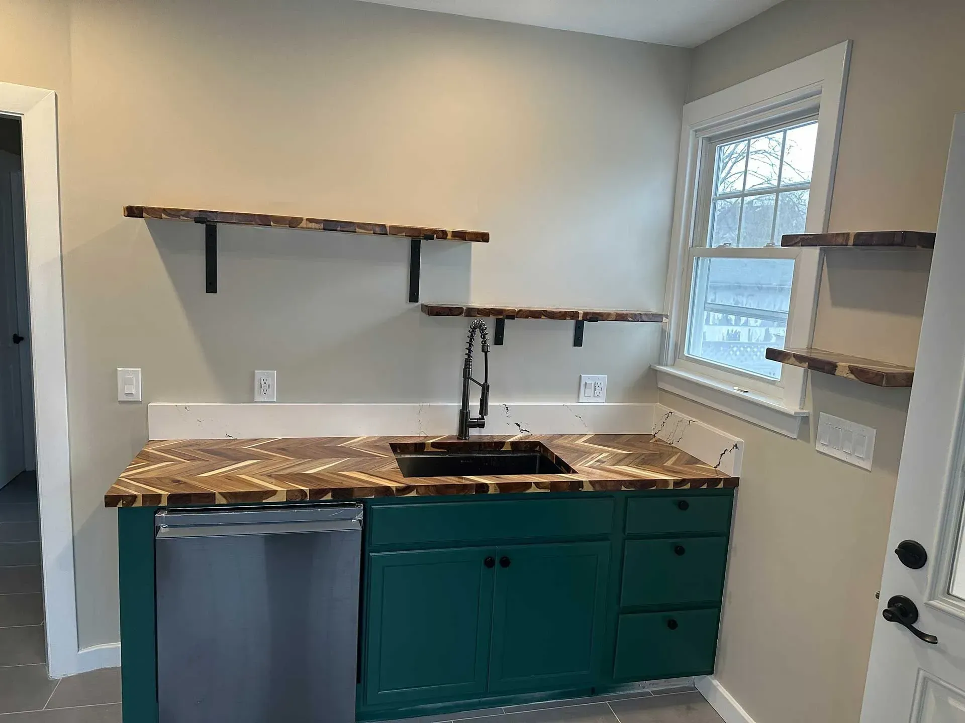 Small kitchen with green cabinets, stainless steel dishwasher, black sink, wood countertops and floating shelves.